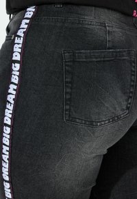 Vaqueros de denim negros con una franja lateral que lleva impreso en letras blancas en negrita "GRANDES SUEÑOS". Incluye un solo bolsillo trasero.