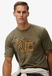 Camiseta de manga corta verde olivo con un gran gráfico de un tigre en amarillo y negro, texto en japonés y un sutil acabado texturizado.