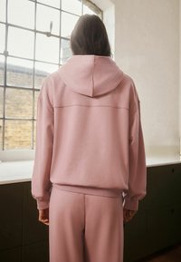 Felpa rosa con vestibilità comoda, dotata di un ampio cappuccio, polsini a coste e una texture morbida. Indossata con pantaloni abbinati, vista da dietro.