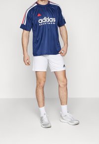 Man i blå Adidas sporttröja, vita Adidas shorts, vita ankelsockor och vita Adidas sneakers som står framför en neutral bakgrund.
