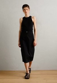 Black sleeveless top, high neckline, paired with a black midi skirt featuring a button detail. Worn with black strappy sandals on wooden floor.