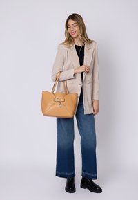 Borsa a mano in pelle color cuoio con forma geometrica e una fibbia decorativa. Il modello indossa un blazer beige, una maglia nera e jeans a gamba larga.
