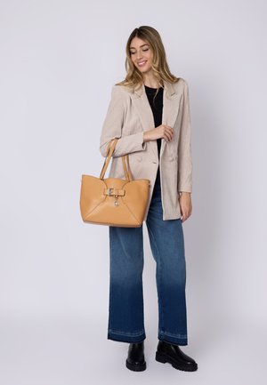 Borsa a mano in pelle color cuoio con forma geometrica e una fibbia decorativa. Il modello indossa un blazer beige, una maglia nera e jeans a gamba larga.