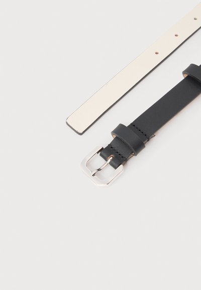 Δύο δερμάτινα λουριά ζώνης, ένα μαύρο με ασημένια αγκράφα και ραφές, και ένα απλό λευκό με διάτρητες τρύπες, τοποθετημένα επίπεδα σε ανοιχτόχρωμη επιφάνεια.