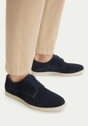 Füße in marineblauen Wildleder-Schnürschuhen kombiniert mit hellbeigen, gekürzten Hosen vor weißem Hintergrund.