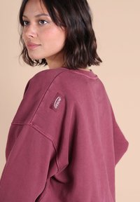 Pull en laine maroon surdimensionné avec une texture douce, comportant un écusson logo sur l'épaule et une coupe décontractée. Fond uni.