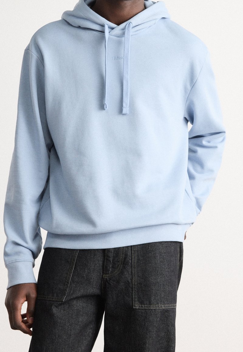 Homme portant un sweat à capuche bleu clair avec cordons et pantalon gris foncé, debout avec une main dans la poche devant un fond uni.