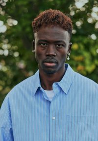 Chemise à manches courtes rayée bleu clair avec un col pointu, portée sur un t-shirt blanc. Petite poche sur le côté gauche, texture lisse.