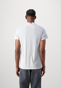 Bílé bavlněné tričko s v-výstřihem, které má malý pruhovaný akcent na límci. Model má na sobě tmavě šedé džíny.