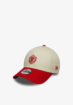Beige und rote Baseballkappe mit Manchester United-Logo vorne und New Era-Emblem an der Seite, aus einem Winkel vor weißem Hintergrund dargestellt.