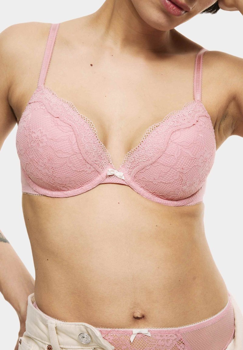 Sostén de encaje rosa con patrón floral, soporte con aros y tirantes ajustables. Detalle de lazo en el centro, complementado por bordes festoneados.