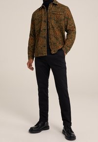 Veste en denim verte et marron à motifs avec deux poches poitrine, portée sur un col roulé noir, assortie à un pantalon slim noir et des chaussures noires.