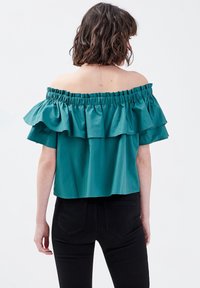 Haut teal décolleté avec des manches volantes, col élastique, coupe ample et texture lisse, associé à un pantalon noir ajusté.