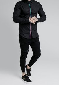 Schwarzes Hemd mit knöpfen und bunten Paspelierungen, tailliertes Design. Kombiniert mit schwarzen zerrissenen Jeans und schwarzen Sneakers mit weißen Sohlen.