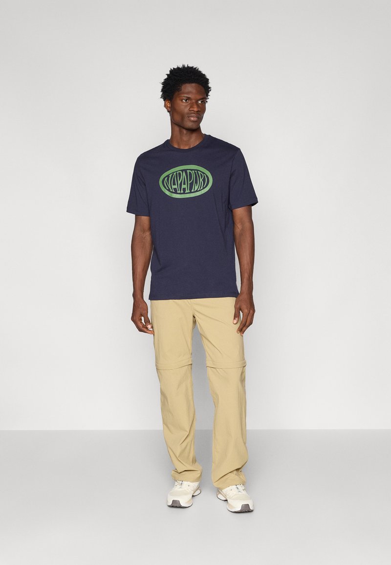 T-shirt bleu marine en coton avec imprimé logo vert, associé à un pantalon beige clair et des baskets blanches. Design simple et décontracté avec des manches courtes.