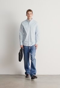 Gaiši zila pogājama krekla, zilas džinsa bikses, turot melnu ādas portfeli, staigā uz gludas pelēkas virsmas baltā fona priekšā.