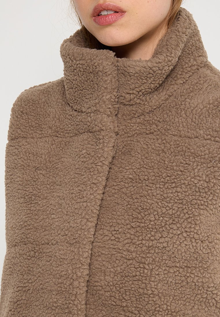 Veste en polaire marron avec col haut, surface texturée, fermetures dissimulées et sans pièces métalliques visibles. Apparence de tissu doux et chaud.