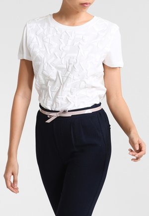 Femme portant une chemise blanche texturée à manches courtes avec des lettres en relief, un pantalon noir et une fine ceinture rose clair nouée en nœud.