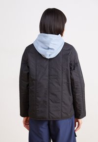 Schwarz gesteppte Jacke mit einer hellblauen Kapuze. Sie hat vertikale Nähte und einen lässigen Schnitt. Getragen über einer marine-blauen Hose, wobei die Rückansicht zu sehen ist.