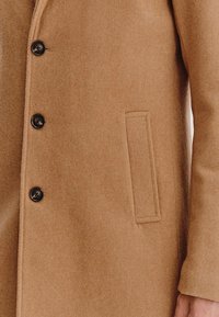 Płaszcz z mieszanki wełny wielbłądziej, charakteryzujący się fakturowanym materiałem, pięcioma czarnymi guzikami oraz boczną kieszenią z prostokątną klapą.