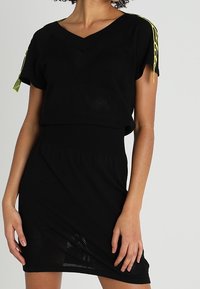 Femme portant une robe en tricot noire avec un décolleté en V et des manches courtes, ornée de détails à cordon jaune fluo sur les épaules.