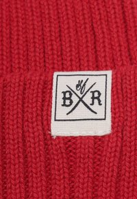 Rød strikket stof med lodrette ribber. Fremtrædende vævet mærke med sort tekst og krydsede symboler på en hvid baggrund.