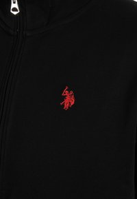 Black zip-up garment with small red embroidered logo of a polo player riding a horse, located on the chest area.