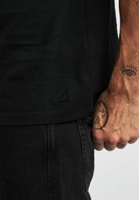 Svart bomullströja, kort ärm, med en texturerad logopatch vid fållen. Bärs med svarta denimbyxor som visar en tatuerad hand.