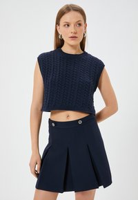 Mörkblå stickad crop top med korta ärmar och en texturerad kabelmönster, tillsammans med en plisserad mörkblå kjol med knappdetaljer.