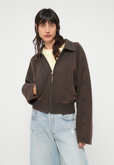 Veste marron courte à fermeture éclair en matière douce avec un grand col et des manches surdimensionnées. Associée à un jean délavé bleu clair et usé.