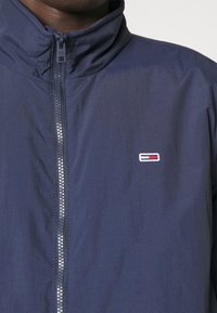 Marineblå jakke med høj krave, frontlynlås og broderet logo med røde og hvide accenter på venstre side. Glat tekstur.