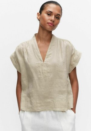 Kvinde med håret i en knold iført en løs beige V-hals kortærmet bluse og hvide bukser, står med hænderne i lommerne mod en hvid baggrund.