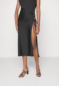 Schwarzer Satin-Midirock mit seitlichem Schlitz, mit Raffung und Bindebändern an der Taille. Kombiniert mit schwarzen Riemchen-Absatzschuhen.