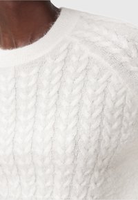 Primo piano di un maglione bianco a treccia indossato da una persona di colore scuro, che mostra la texture e i dettagli della cucitura vicino al colletto.