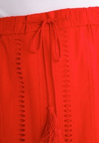 Jupe en tissu rouge avec une taille à cordon, des motifs découpés complexes et des franges décoratives à l'avant.