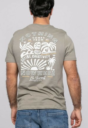 Homme portant un T-shirt gris-vert avec un motif tropical blanc et jaune et les inscriptions « Nothing comes from nowhere » et « Be Yourself », jean bleu.