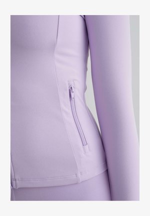 Top de manga larga lila claro hecho de un material suave y elástico, con un bolsillo lateral con cremallera y detalles de costura limpios.