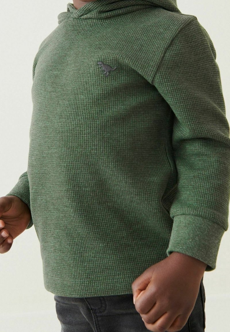 Enfant portant un sweat à capuche vert texturé à manches longues avec un petit emblème de dinosaure sur la poitrine, debout les bras légèrement pliés.