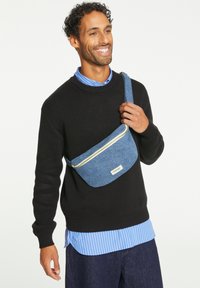 Sac en denim bleu porté en bandoulière avec un design plat, fermeture éclair jaune et une étiquette en tissu. Porté par-dessus un pull noir et une chemise à rayures bleues.