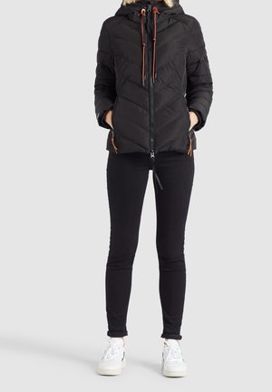 Persona con chaqueta acolchada negra con capucha con borde de piel, pantalones ajustados negros, zapatillas blancas, con las manos en los bolsillos de la chaqueta, de pie sobre un fondo liso.