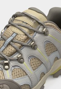 Μπεζ και γκρι πλεκτό παπούτσι πεζοπορίας με γκρι κορδόνια, μεταλλικά οπές και το λογότυπο της «The North Face» στη γλώσσα, παρουσιάζοντας λεπτομερή κοντινή εικόνα.