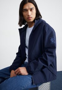 Veste légère navy avec capuche, poignets élastiques et zip frontal. Dotée d'une patch logo Timberland sur le bras gauche. Coupe décontractée.
