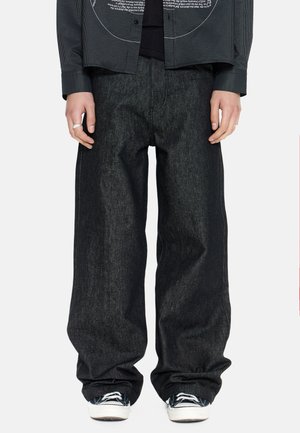 Persona con pantalones de mezclilla negros holgados combinados con zapatillas negras con suelas blancas y cordones negros, manos relajadas a los lados.