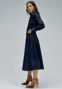 Vestido de mezclilla, azul oscuro, con cuello abotonado, mangas largas, bolsillos laterales, falda en línea A, acentuado con costuras, combinado con zapatos de tacón marrones.