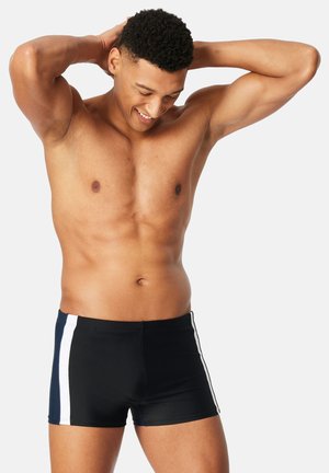 Uomo atletico che indossa pantaloncini da bagno neri con strisce bianche ai lati, sorridente, con le mani dietro la testa, su uno sfondo semplice.