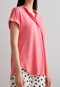 Rosa Kurzarm-Pullover mit Henley-Ausschnitt. Weiches, leichtes Material mit einer lockeren Passform und abgerundetem Saum. Glatte Textur.