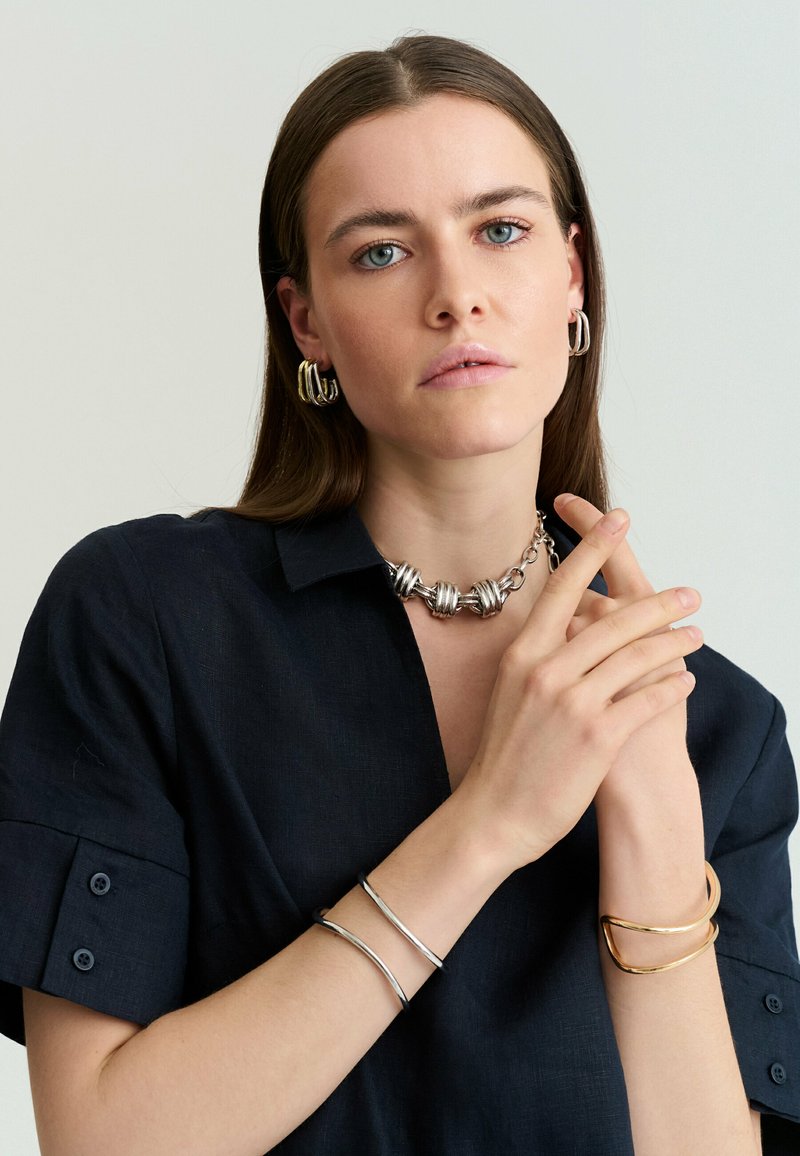 Femme aux cheveux bruns raides portant une chemise sombre, des boucles d’oreilles en argent, un collier argent massif et des bracelets en métal mélangé aux deux poignets.