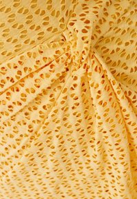 Gul, strukturerad tyg med ett perforerat mönster och små öppningar. Tyget är samlat på ena sidan med en twistdetalj.