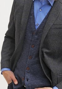 Mörkgrå blazer över en blå knapp-up skjorta och en marinblå kavaj med bruna knappar, som har en strukturerad tyg och skräddarsydd passform.