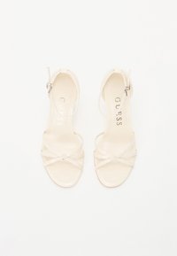 Beige open-toe heeled sandals with crossed straps, thin ankle strap, and a minimalist design. Logo printed on the insole. Smooth texture.